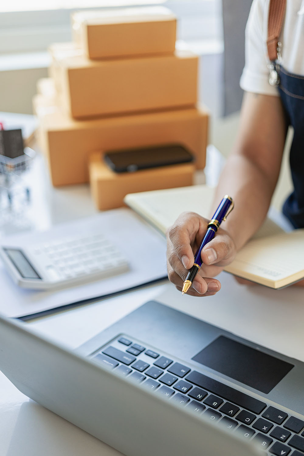 Business owner working on laptop near a notepad, calculator, and boxes on a desk.