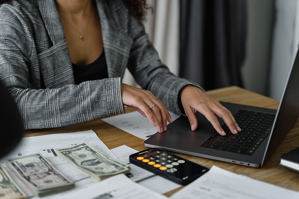 A woman using a laptop near a calculator, paperwork, and cash on a desk.