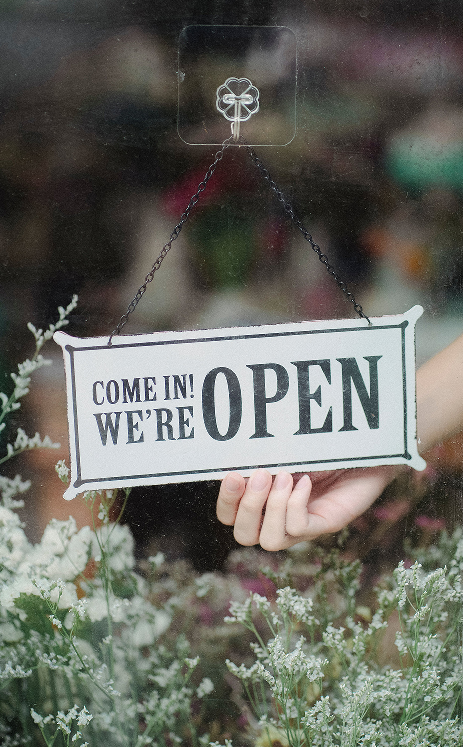 A hand inside a flower shop turning over a sign that reads, "Come in! We're open"