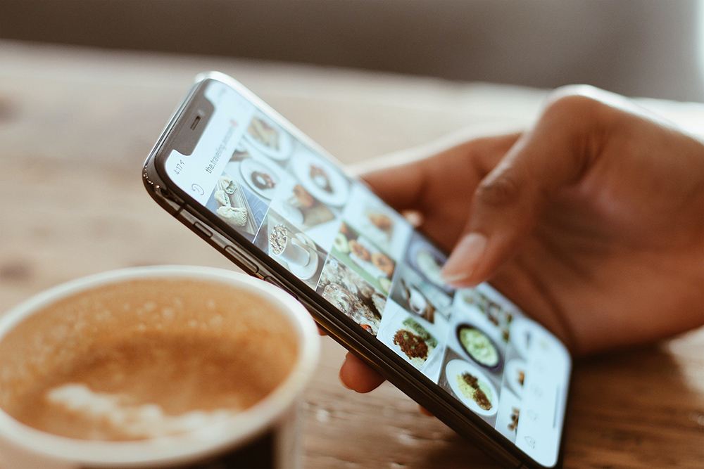 A hand holding a cell phone over a cup of coffee on a table.