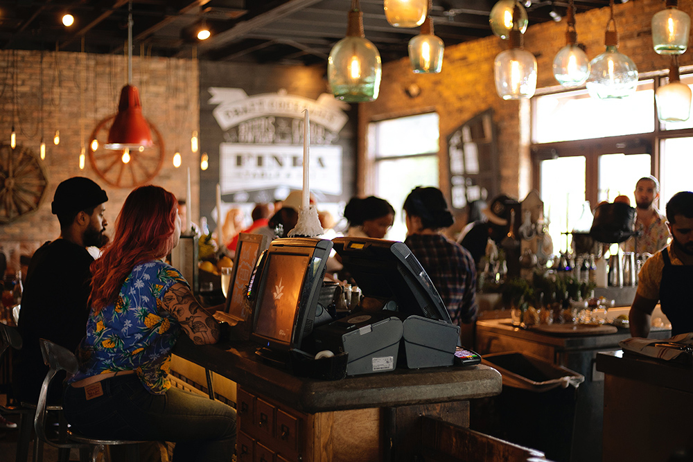 A restaurant setting with people working behind a bar and sitting at tables with point of sale systems in the foreground.