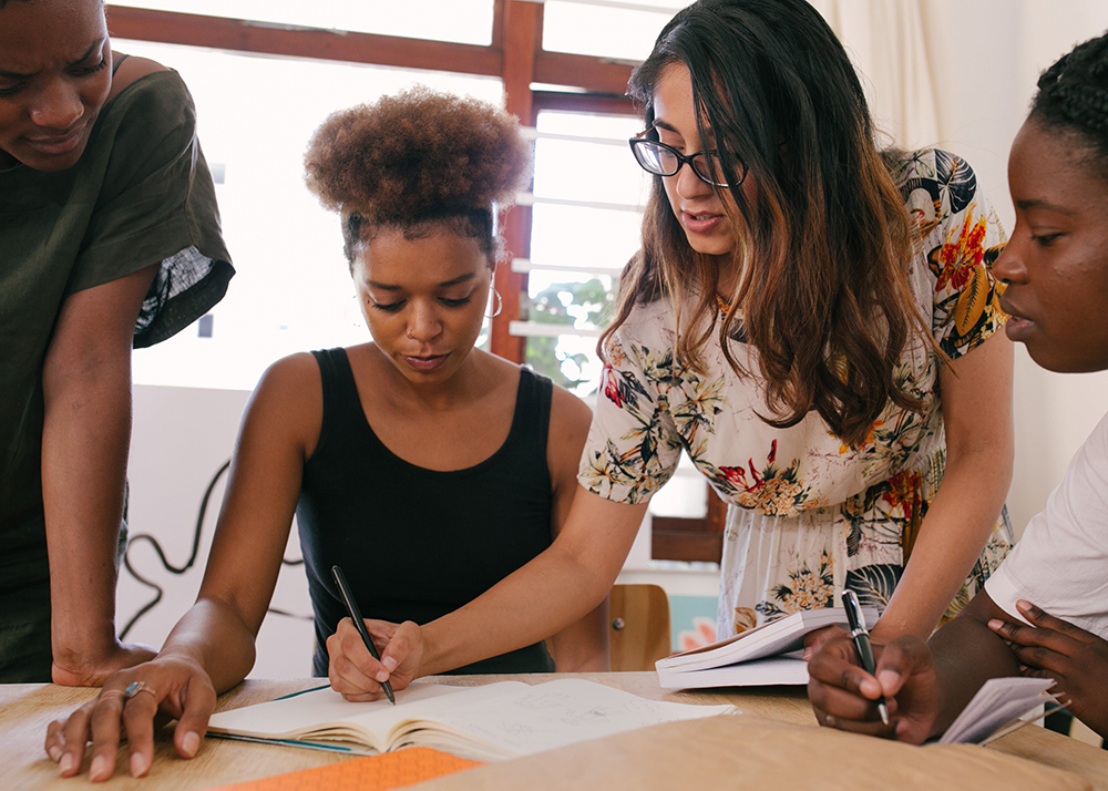 Three people looking on as one person writes on a piece of paper on a table.