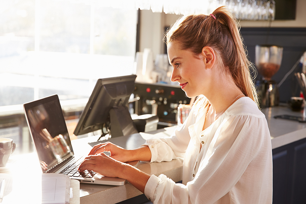 A woman sits at a desk with a laptop at a restaurant bar.