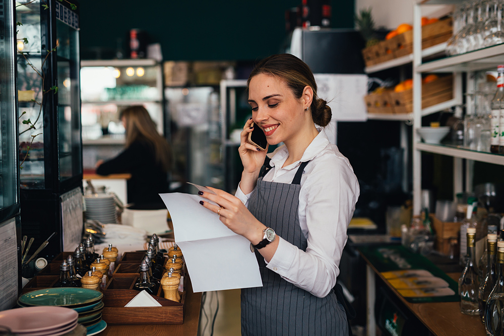 A woman holds a phone to her ear while reviewing a piece of paper in a restaurant environment.