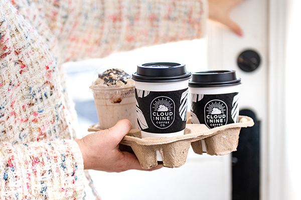 A person's hand carrying a bagasse cup carrier full of hot and cold coffees moves towards the door being opened by the other hand.