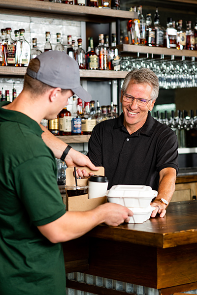 A customer in a ball cap picks up their order from a person behind a bar.