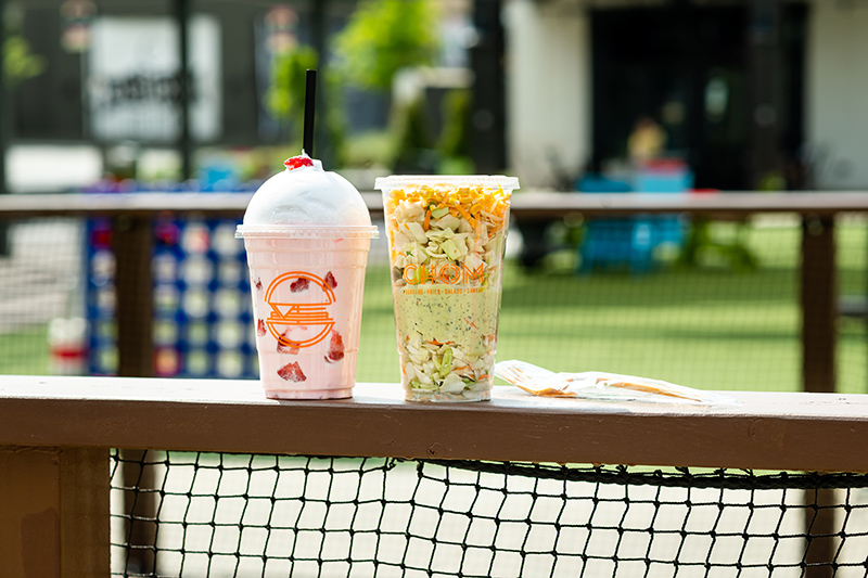 Two custom printed plastic cups on a wooden bar. One has a pink milkshake with whipped cream, while the other has a salad to-go.